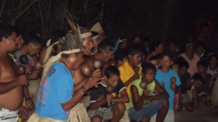 Fotografia noturna de um grupo com cerca de 30 pessoas reunidas num ambiente de terra arenosa e rodeado por árvores com poucas folhas. Todos são indígenas e estão em semicírculos. De perto, são vistos homens e meninos, alguns deles usam camisa e bermuda, outros estão de peito nu. Alguns estão com cocar na cabeça e usam saia de palha. A maioria segura um maracá com as mãos.