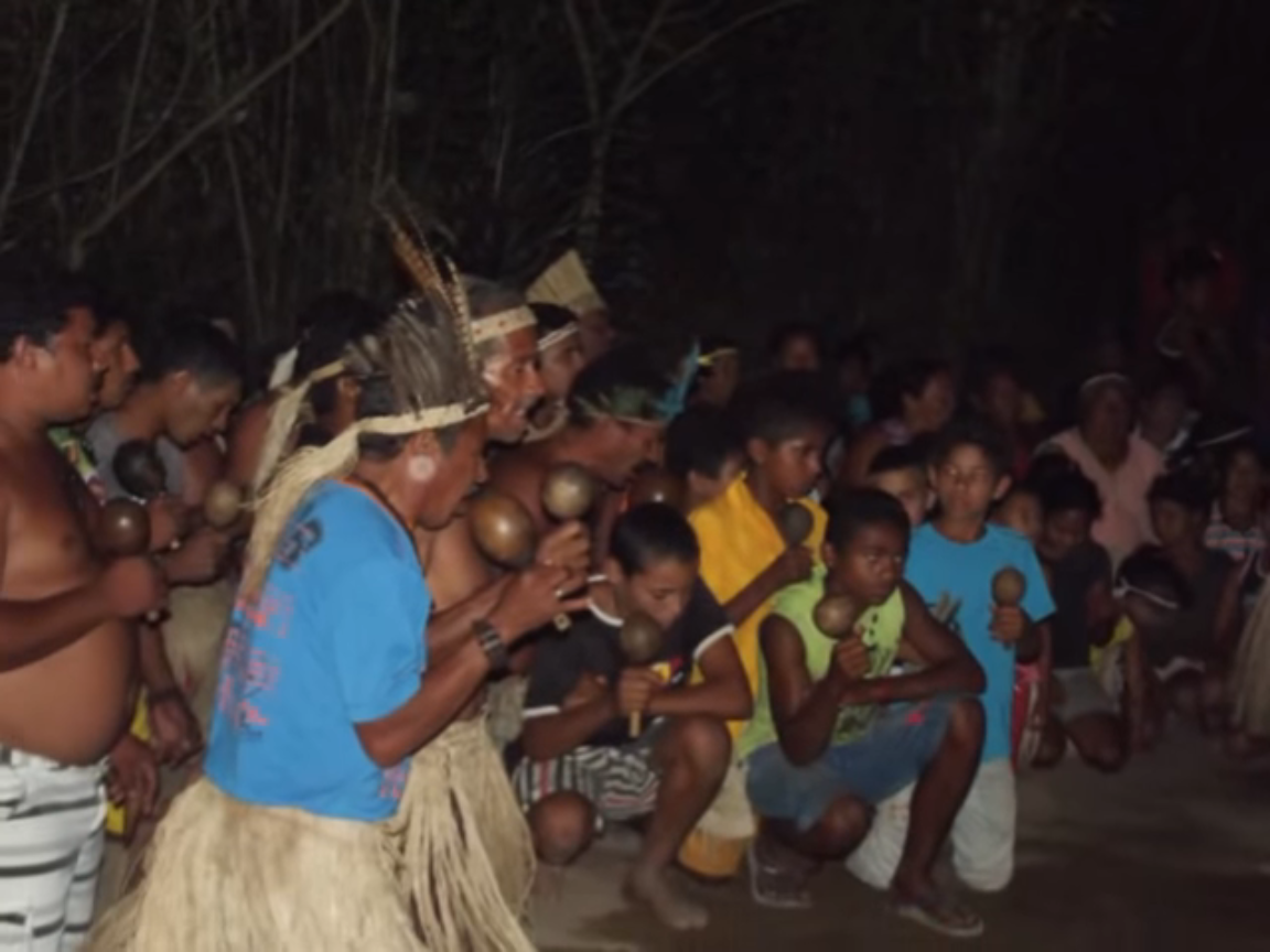 Fotografia noturna de um grupo com cerca de 30 pessoas reunidas num ambiente de terra arenosa e rodeado por árvores com poucas folhas. Todos são indígenas e estão em semicírculos. De perto, são vistos homens e meninos, alguns deles usam camisa e bermuda, outros estão de peito nu. Alguns estão com cocar na cabeça e usam saia de palha. A maioria segura um maracá com as mãos.