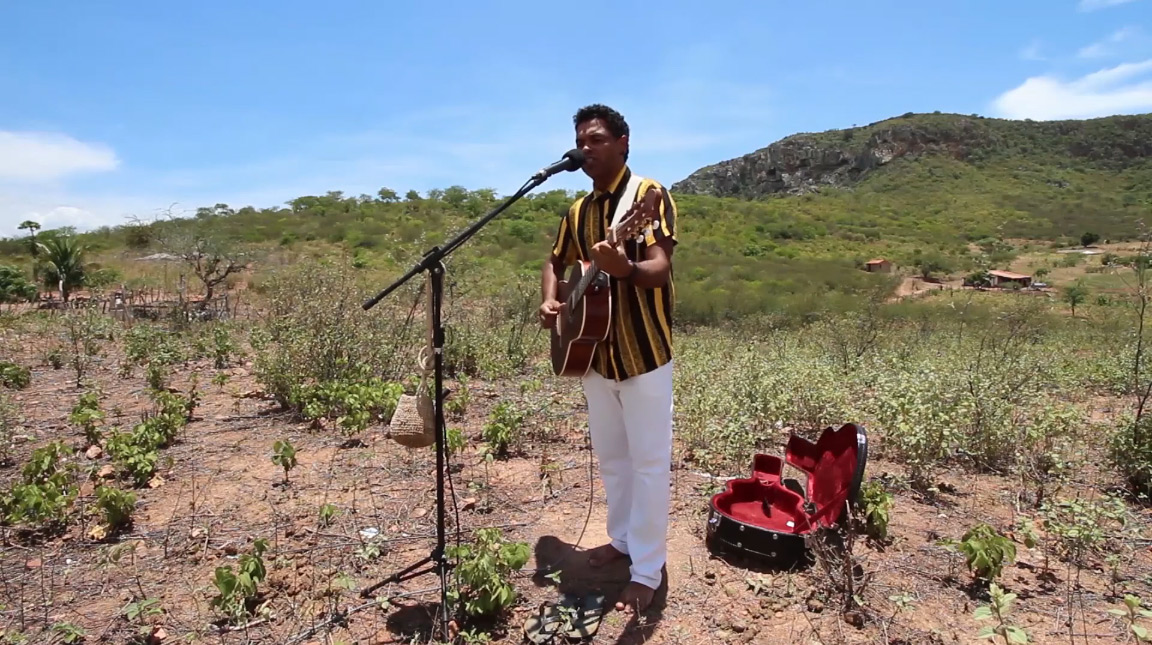 Fotografia paisagem de um homem indígena tocando violão numa área de campo, com vegetação rasteira ao redor. Ele está em pé, com um cavalete e microfone à sua frente e caixa do violão aberta no chão. O homem tem pele escura e cabelos pretos curtos. Usa uma camisa de botão listrada com tons marrons e calça branca. Ao fundo, céu claro, com algumas nuvens e colinas.