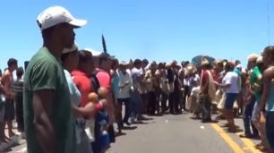 Fotografia de um grupo indígena reunidos em uma BR, sob o sol que ilumina o grupo. Os homens e as mulheres estão em pé, bem próximos uma das outras, e juntas se apresentam em um formato circular. De perto, eles usam camisa e bermuda e alguns seguram maracá. Ao longe, mais indígenas com cocar na cabeça e saia de palha. No acostamento esquerdo, outras pessoas.