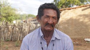 Fotografia de um homem indígena de frente, visto do tórax para cima. Ele aparenta ter mais do que 55 anos, tem a pele escura, cabelos e bigodes pretos. Os olhos são puxados, nariz largo e boca fina Ele está sério, com o olhar apertado, sobrancelhas e boca franzidas, destacando algumas linhas de expressões na testa e ao redor dos olhos. Ele usa camisa de botão e colar de sementes. No fundo, o chão é arenoso alaranjado, há uma cerca de madeira, uma casa de barro, árvores e uma forte luz do sol.