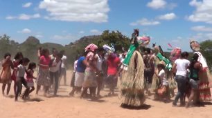 Fotografia em paisagem de grupo com cerca de 20 mulheres e meninas indígenas que caminham da esquerda para direita, formadas em trio ou dupla, sob a luz do sol. Entre elas, três cobertas por um manto de palha da cabeça aos pés, com adereços coloridos no topo. Outras usam blusa e saia. Ao longe, uma montanha com vegetação rasteira e algumas árvores, sob o céu azul com algumas nuvens brancas.