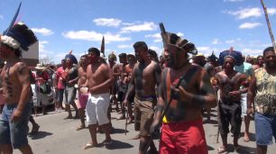 Fotografia de um grupo de homens indígenas que caminham da direita para esquerda no asfalto, sob a luz do sol. Muitos deles estão com cocar na cabeça, peitoral nu e corpos pintados com tinta de jenipapo e orucum, nas cores preta e vermelha. Alguns seguram pedaços de pau e as bocas estão semiabertas.