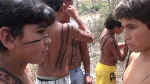 Fotografia paisagem ao ar livre de seis crianças indígenas com o peitoral nu, com cerca de 10 anos. De perto, um menino de pele parda e outro de pele clara, frente a frente, são vistos de perfil direito. Ambos estão com semblante sério. O de pele parda está com tinta preta de jenipapo no rosto, ele olha de lado. O de pele clara está com olhar vidrado para frente e lábios cerrados. Um pouco atrás, as crianças estão em dupla e se pintam com traços e bolinhas. Há uma criança que é vista apenas uma pequena parte do seu braço.