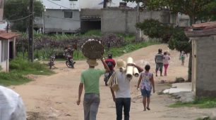 Fotografia de uma rua de terra alaranjada, com várias pessoas indígenas de costas, em caminhada. Em destaque, um homem de camisa verde que carrega um fardo de palha na cabeça. Ao lado, outro homem que carrega tubos de papelão. Mais adiante, uma mulher e um homem em uma motocicleta. A rua é ladeada por pequenas casas de alvenaria, vegetação rasteira e algumas árvores.