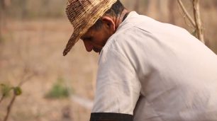 Fotografia de um homem indígena de perfil esquerdo, inclinado para baixo. Ele é uma pessoa mais velha, usa um chapéu de palha e uma camisa branca de manga longa. O chapéu cobre uma parte de seu rosto e o seu olhar está fixo para o chão. O fundo é desfocado com vegetação seca.