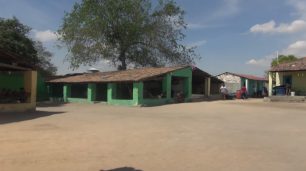 Fotografia de uma área de campo, com extenso chão de terra batida alaranjado. Mais distante, casas pequenas com paredes verdes ou amarelas e telhados de telhas. Os terraços são abertos, com colunas quadradas. Um deles comporta uma caixa d’água no chão. Por trás da casa verde, há uma enorme árvore e ao redor, transeuntes e motocicletas estacionadas.