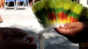 Fotografia paisagem de duas mãos que segura um cocar de penas coloridas: verde, amarelo e vermelho. Do lado esquerdo, uma das mãos segura a base, feito por uma linha grossa trançada. Enquanto, do lado direito, a outra mão segura as penas. No fundo, é visto o chão e algumas cadeiras de metal.