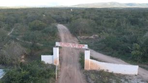 Fotografia da vista aérea de uma estrada de terra ladeada por vegetação rasteira e densa. No centro, há uma entrada com portão aberto e muro branco nas laterais, com um letreiro na cor vermelha no topo: Tribo Indígena Fulni-ô Ouricuri. Proibido a entrada. Não se aproxime". Ao fundo, bem distante, há montanhas e o céu está claro.