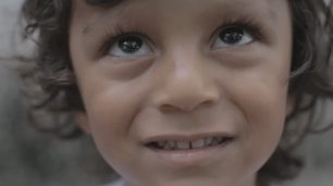 Fotografia do rosto de uma criança indígena. É um menino, de pele parda, cabelos ondulados castanho-escuros e olhos grandes e brilhantes. As pupilas dos seus olhos estão levemente viradas para cima e ele sorri de modo sutil.